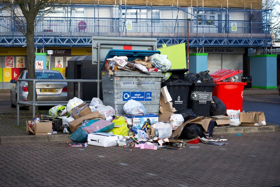 A pile of mixed rubbish, including black plastic trash bags, a yellow plastic storage container, a worn and dirty beige car seat, and miscellaneous waste items, is situated on a gravel surface next to